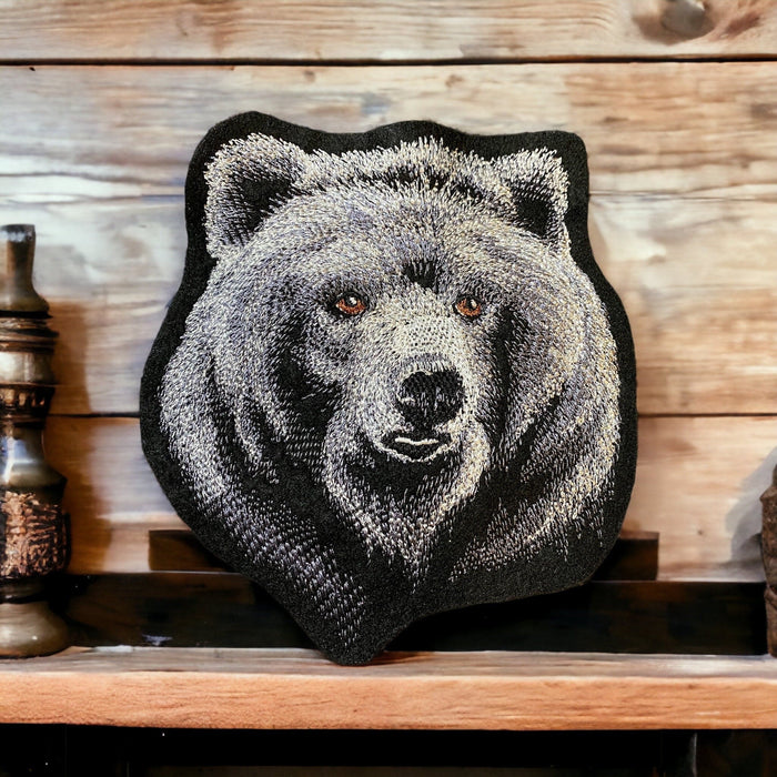 Embroidered patch shaped like a bear’s head, featuring detailed gray and brown stitching, displayed on a rustic wooden shelf.