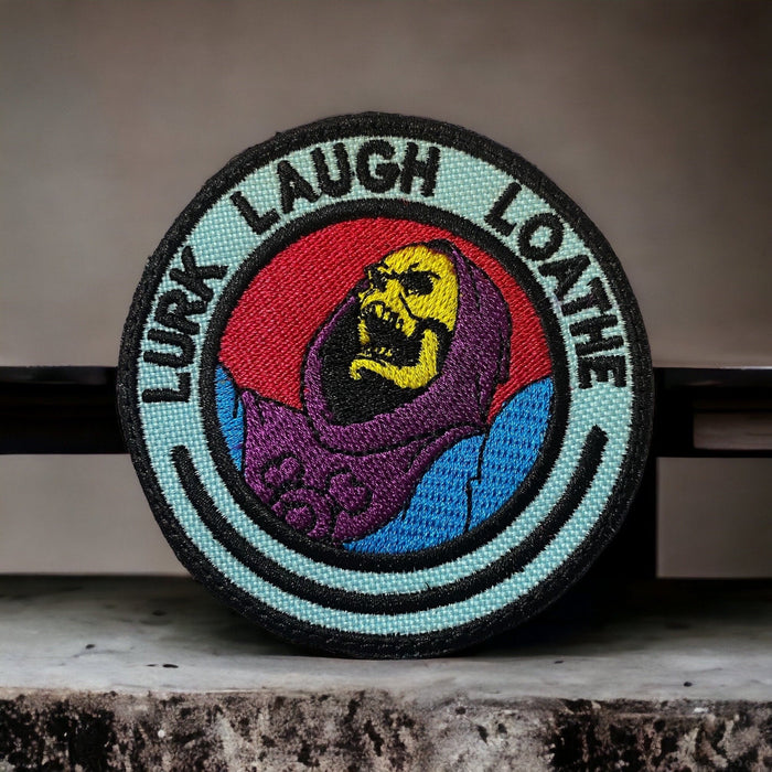 Round embroidered patch showing a cartoon skull-faced villain with the text ‘Lurk Laugh Loathe.